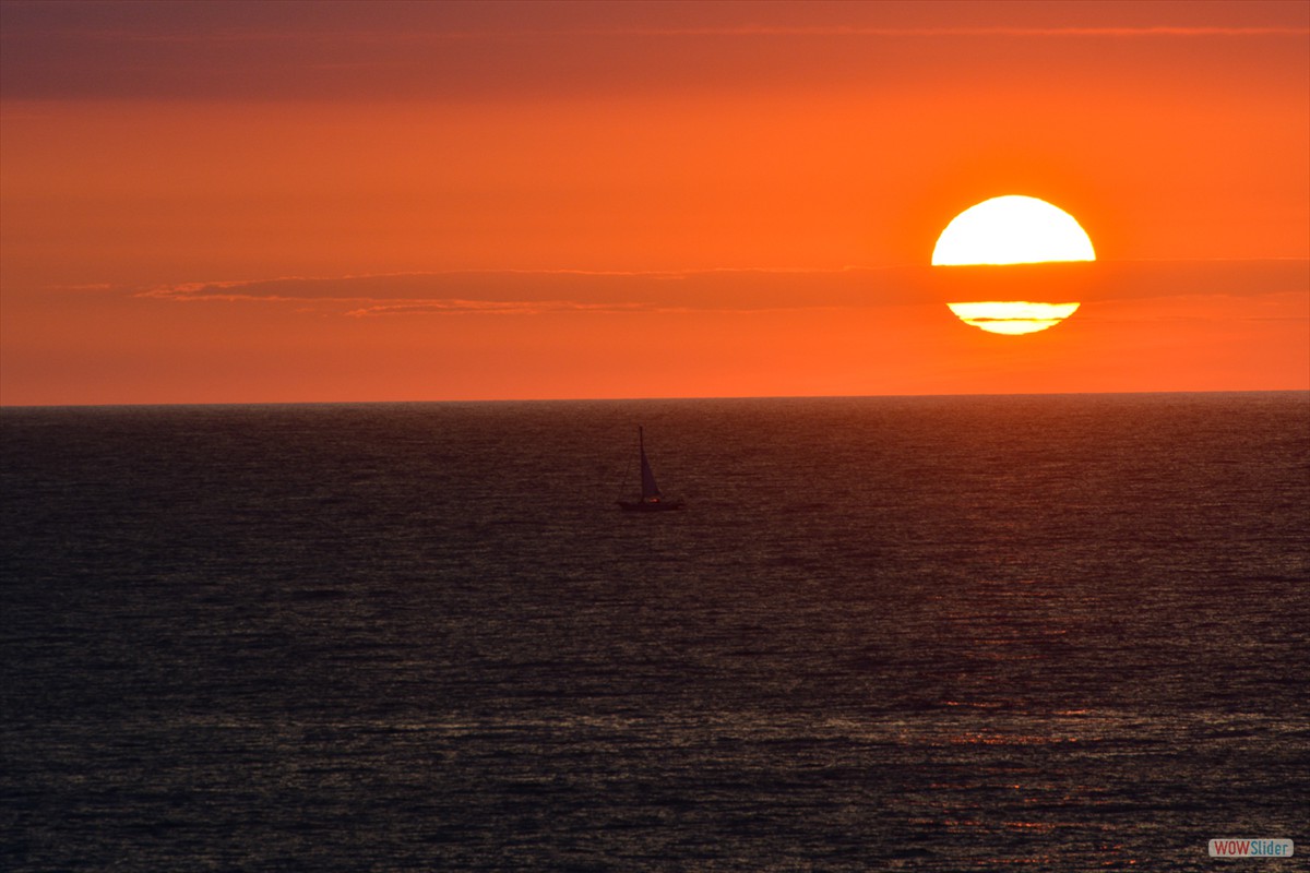 A large fireball paints an orange glow across the sky using a zoom lens as a sailboat passes by.